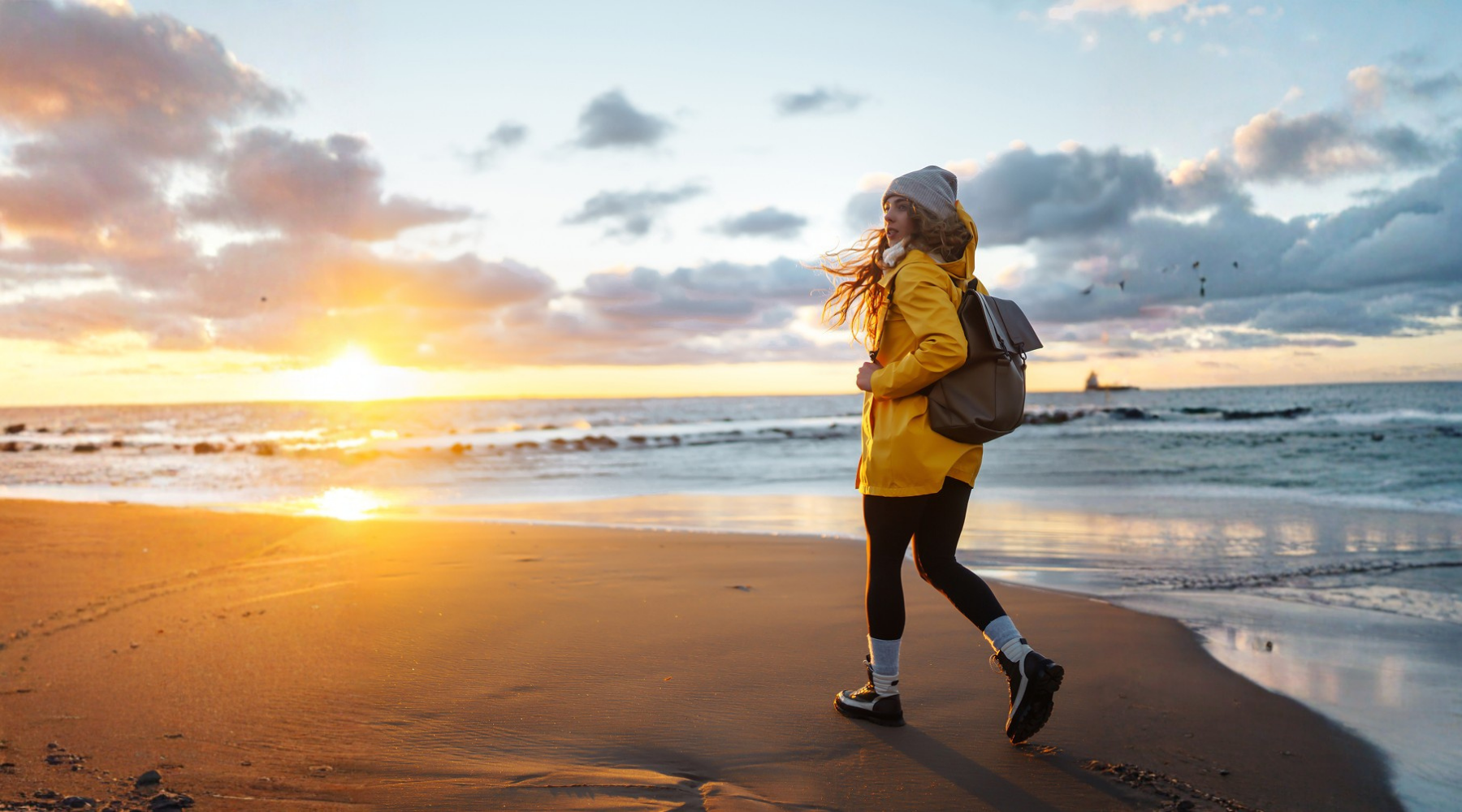 Teen girl walking along a beach at sunrise in a yellow raincoat and backpack, illustrating gentle morning movement and outdoor recovery for adolescents during winter.