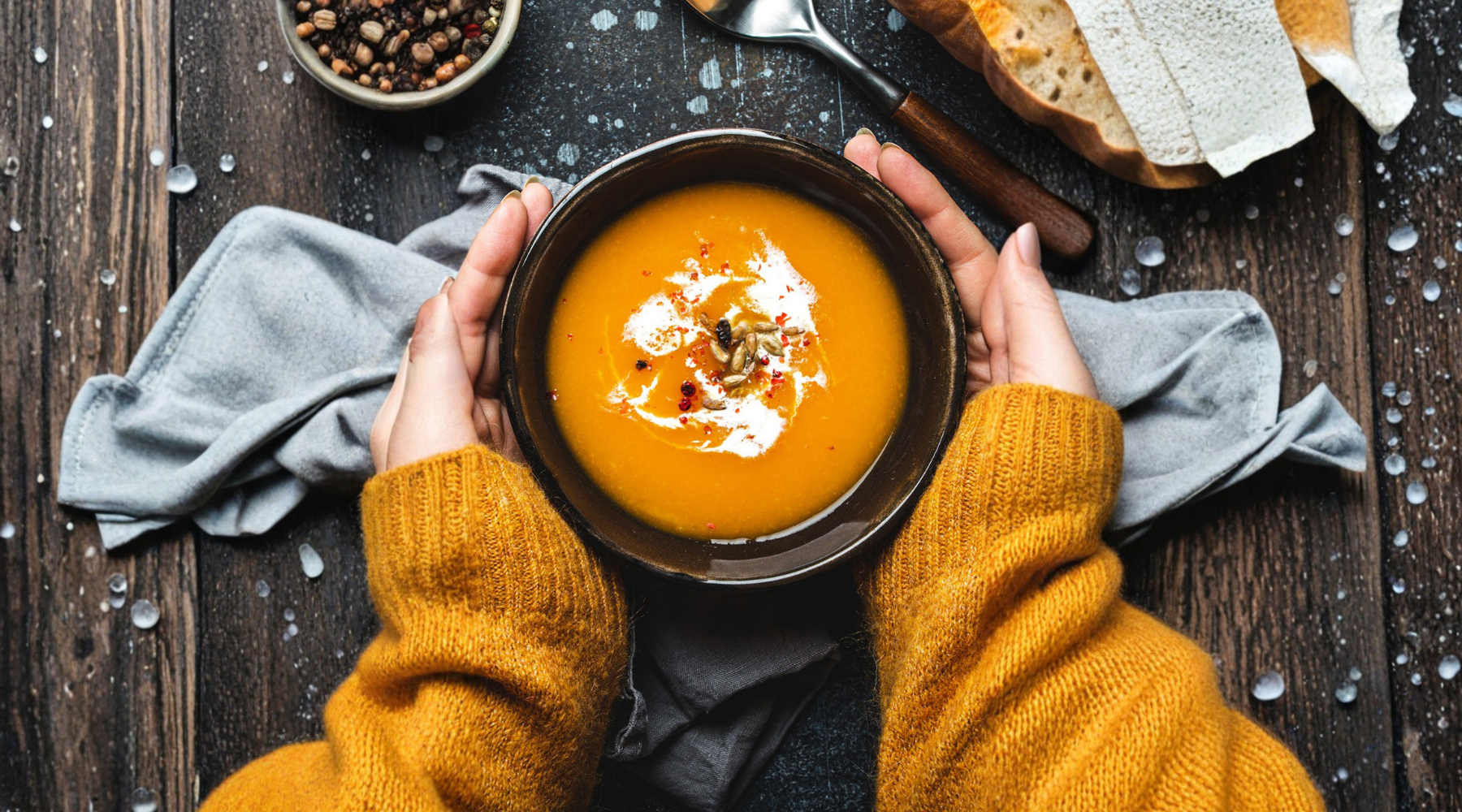 Hands in a mustard knit jumper cradling a warm bowl of pumpkin soup topped with cream and seeds, illustrating nourishing whole food nutrition for teen recovery during winter.